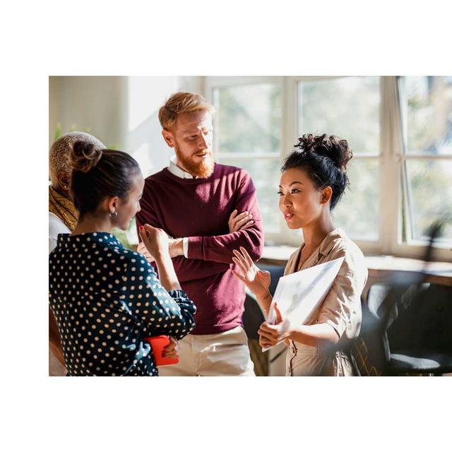 A group of diverse office workers listen intently as one woman speaks to them, referencing a tablet.