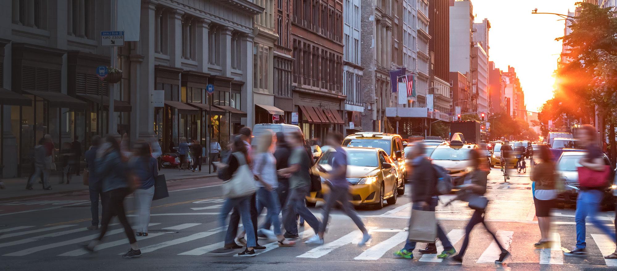 An image showing a vibrant community. A bright sunset in the background and people crossing a busy street at a crosswalk in the foreground.