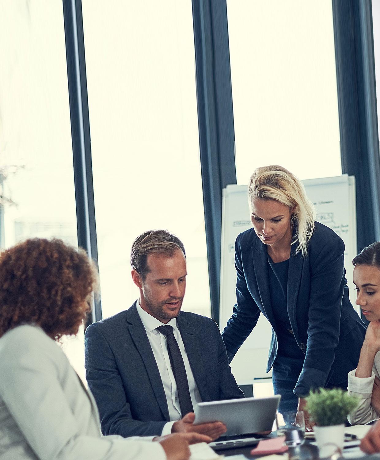 A diverse group of office workers consult a laptop screen - some are seated and others are standing, all appearing very concentrated.